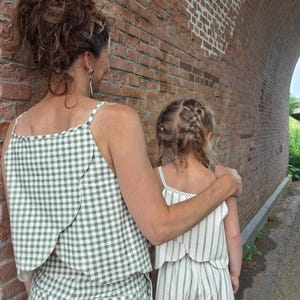 Op de afbeelding: Een mouwloze top met saliegroen en wit vichy-ruitpatroon en bijpassende broek gedragen door een volwassene, naast een kind in een witte mouwloze top met verticale saliegroene strepen en bijpassende broek, beide met een gelaagde schulprand.