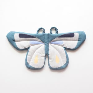 May include: A butterfly-shaped textile accessory in shades of teal, white, and navy blue. The wings feature light blue and yellow teardrop-shaped accents. Two small, gathered fabric loops are attached to the top center of the butterfly wings.