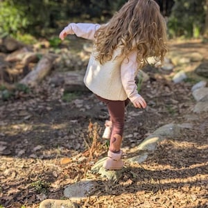 May include: A young person with long curly brown hair is wearing a white fleece vest, a pink long-sleeved shirt, brown leggings, and brown boots. They are standing on a path made of wood chips and rocks.