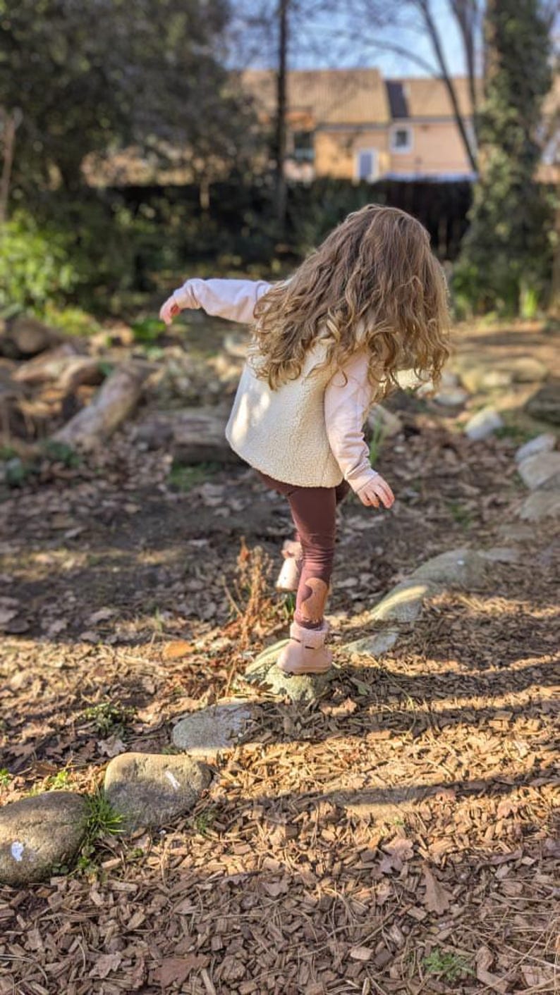May include: A young person with long curly brown hair is wearing a white fleece vest, a pink long-sleeved shirt, brown leggings, and brown boots. They are standing on a path made of wood chips and rocks.