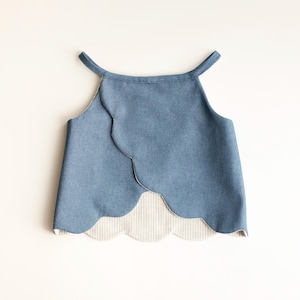 Op de afbeelding: Een blauwe denim top met een geschulpte zoom en halslijn. De top heeft een halter-stijl ontwerp met bandjes en een contrasterende gestreepte stof aan de binnenzoom. Het kledingstuk ligt plat op een wit oppervlak.