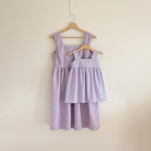 Puede incluir: Dos vestidos lavanda a juego colgando de perchas de madera. El vestido más grande tiene un escote cuadrado y una falda fruncida. El vestido más pequeño tiene un corpiño a rayas y una falda fruncida. Ambos vestidos están hechos de tela ligera.