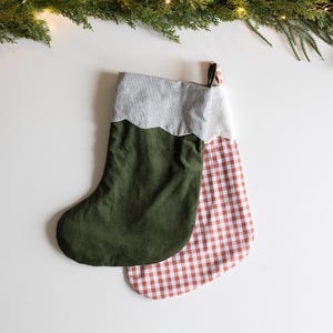 May include: Two Christmas stockings. One is olive green with a scalloped edge and a striped top. The other is red and white gingham. The stockings are on a white surface with evergreen branches and lights in the background.