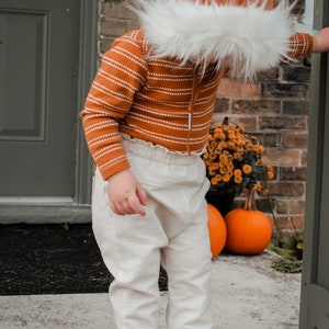May include: A child wearing a brown and white striped sweater, white pants, and brown shoes. The child is wearing a brown hooded hat with a white fur trim. The child is standing in front of a brick wall with pumpkins and flowers.