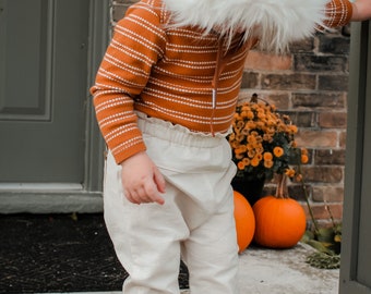 May include: A child wearing a brown and white striped sweater, white pants, and brown shoes. The child is wearing a brown hooded hat with a white fur trim. The child is standing in front of a brick wall with pumpkins and flowers.
