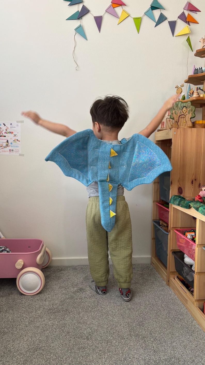 K&ouml;nnte beinhalten: Ein Kind tr&auml;gt ein hellblaues Drachenkost&uuml;m mit gelben Akzenten. Das Kost&uuml;m hat gro&szlig;e Fl&uuml;gel und einen Schwanz. Das Kind steht in einem Raum mit einem bunten Banner und Holzregalen.