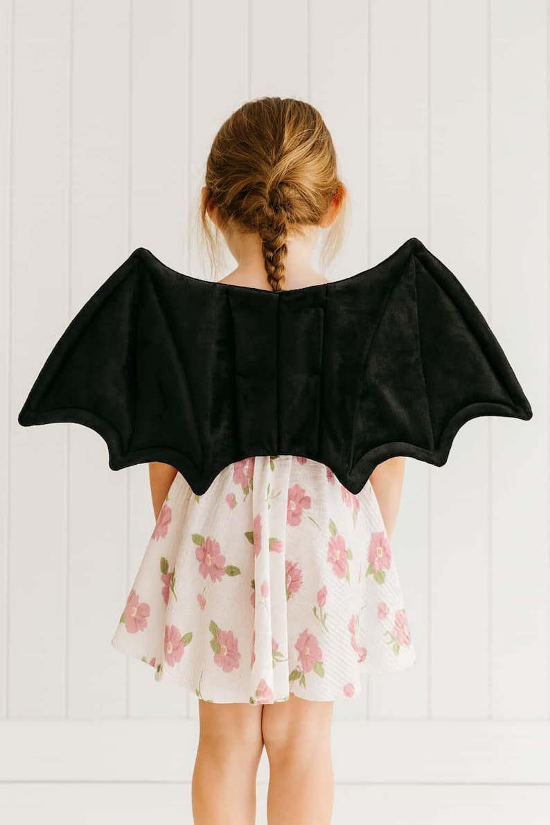 K&ouml;nnte beinhalten: Ein Kind tr&auml;gt ein schwarzes Fledermausfl&uuml;gel-Kost&uuml;m. Die Fl&uuml;gel bestehen aus einem weichen, samtigen Material und sind am R&uuml;cken befestigt. Das Kind tr&auml;gt ein wei&szlig;es Kleid mit rosa Blumenmustern.