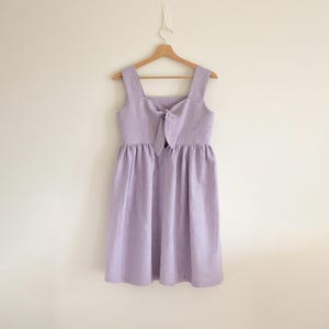 Puede incluir: Un vestido de lino lila con escote cuadrado y detalle de lazo en el busto. El vestido tiene cintura fruncida y llega a la rodilla. Cuelga de una percha de madera sobre un fondo blanco liso.