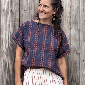 Op de afbeelding: Een marineblauw shirt met korte mouwen, voorzien van verticale rijen kleine oranje bloemmotieven en dunne oranje strepen, gecombineerd met een witte broek met verticale rode strepen. Het shirt heeft een brede boothals en een losse pasvorm.