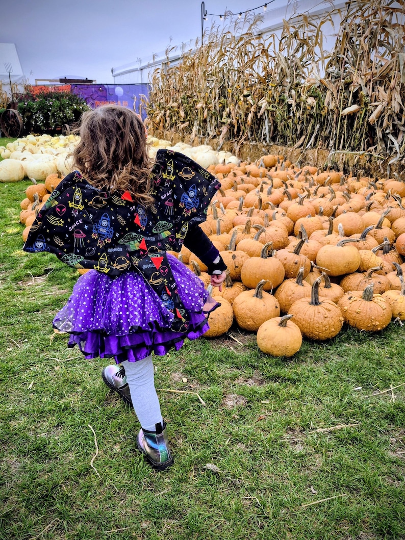K&ouml;nnte beinhalten: Ein Kind mit einem schwarzen Umhang mit Weltraum-Design, einem lila Tutu und silbernen Stiefeln rennt durch ein Feld mit K&uuml;rbissen. Die K&uuml;rbisse sind orange und unterschiedlich gro&szlig;, mit Maisstengeln im Hintergrund.