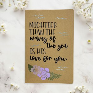 May include: A rectangular kraft paper notebook featuring the black printed text MIGHTIER THAN THE WAVES OF THE SEA IS HIS LOVE FOR YOU. The cover is decorated with light blue wave illustrations and a cluster of purple flowers and green leaves.
