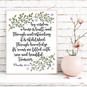 May include: A framed print with a quote from Proverbs 24:3-4, featuring black script and floral accents in green, pink, and blue. The print is displayed on a white shelf with a pink vase holding flowers.