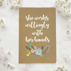 May include: A rectangular notebook with a tan kraft paper cover featuring the white text "she works willingly with her hands" in a script font, positioned above a colorful floral illustration with pink, blue, and yellow flowers.