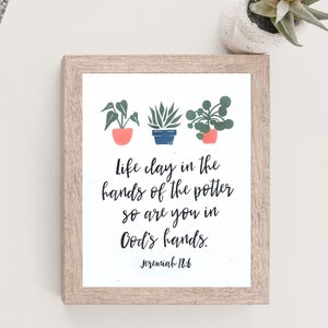 May include: A framed print with a white background and a quote about life being like clay in the potter's hands. The quote is from Jeremiah 18:6. The print features three potted plants with green leaves and pink and blue pots.