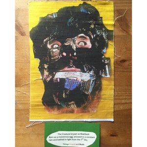 May include: A yellow fabric patch with a black and white collage of a face. The face has a black eye and a blue and purple eye. The text on the patch reads "The Creature known as Bearback Born as a monstrous egg, encased in a constant rain and bathed in light from the 7th Sky. Things Found and Made".