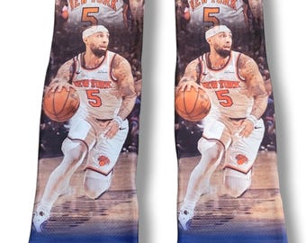 May include: A pair of crew socks featuring a photographic print of a basketball player in a white New York jersey with the number 5, dribbling a basketball. The bottom section of each sock displays the blue and orange New York Knicks team logo.