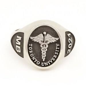 May include: Silver ring with a dark oval centre featuring the Toronto University logo and the text "TORONTO UNIVERSITY". The ring also has the text "MB" and "2021" on the sides. The ring has a medical caduceus symbol.