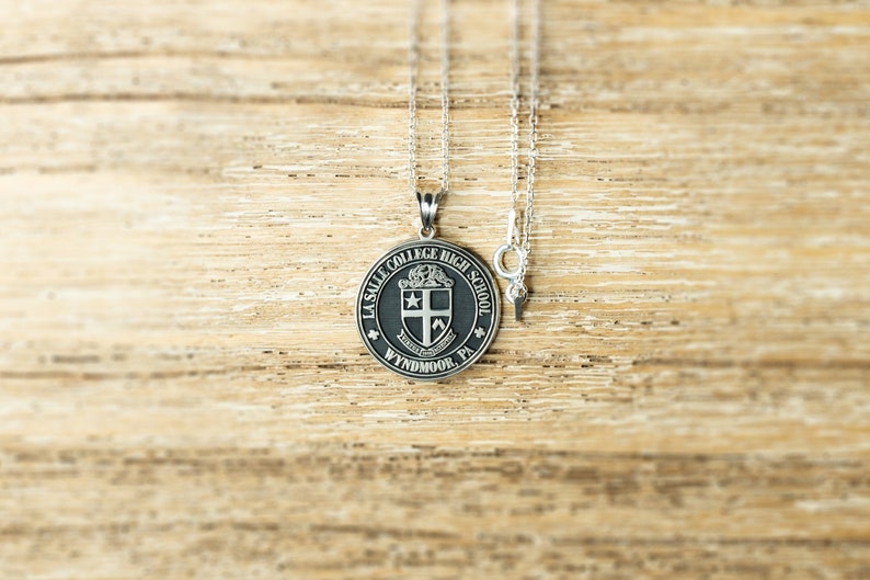 May include: A silver chain necklace with a round pendant featuring the La Salle College High School crest. The crest is black with a silver shield and the words "La Salle College High School" and "Wyndmoor, PA" around the edge.