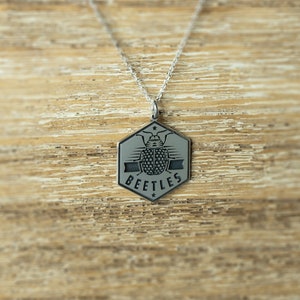 May include: A silver necklace with a hexagonal pendant featuring a stylized beetle design and the word "BEETLES" inscribed on it.