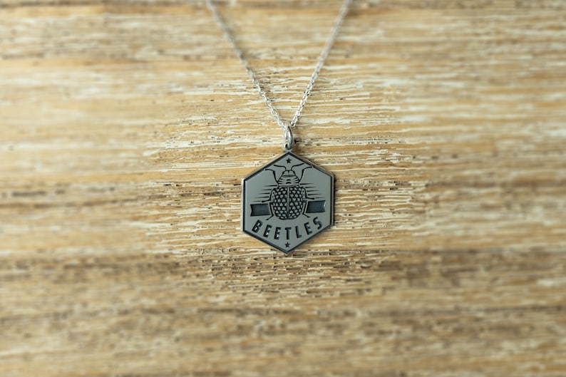 May include: A silver necklace with a hexagonal pendant featuring a stylized beetle design and the word "BEETLES" inscribed on it.
