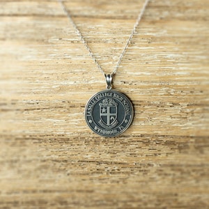 May include: Silver pendant necklace with a round black and white La Salle College High School crest. The crest features a shield with a cross and a star. The text around the crest reads "La Salle College High School, Wyndmoor, PA."