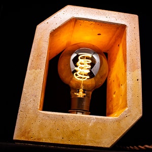 May include: A concrete lamp with a warm, glowing light bulb inside. The lamp is made of a rough, textured material and has a rectangular shape.