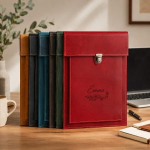 May include: A collection of five vertical leather document folders in mustard yellow, black, blue, dark brown, and red. The red folder features a silver metal clasp, a flap closure, and the name Emma engraved with a floral design on the front.