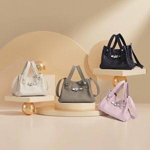 May include: Four leather handbags in various colours: off-white, grey, black, and lavender. Each bag features a top handle, a silver-tone clasp, and a structured design. The bags are displayed on pedestals with gold accents, highlighting their elegant style.