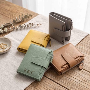 Peut inclure: Quatre portefeuilles en cuir de couleurs jaune, gris, vert et marron. Chaque portefeuille est doté d'une fermeture à bouton-pression et d'une fermeture éclair. Le portefeuille vert porte les initiales "K&C". Présentation sur une surface en bois.
