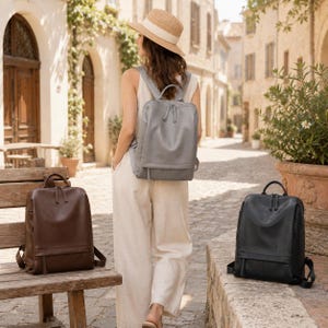 May include: Three leather backpacks in grey, brown, and black. The grey backpack is worn on the back, while the brown one sits on a wooden bench and the black one rests on a stone ledge. Each bag features a main compartment and a front pocket.