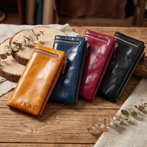 May include: Four leather wallets in shades of tan, blue, burgundy, and black, each with a gold zipper. The tan wallet has "K & C" embossed on the front. Wallets are arranged on a wooden surface.