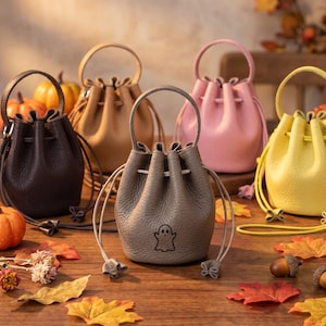 May include: A collection of five small, round, drawstring bucket bags in dark brown, tan, gray, pink, and yellow. The gray bag features a small, black, line-art ghost icon on the front. Each bag has a single top handle and a drawstring closure.