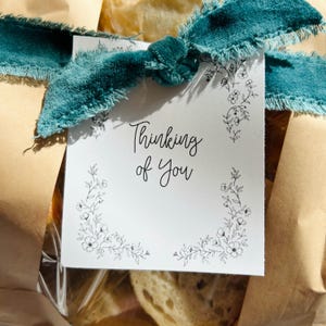Può includere: Un regalo avvolto in carta marrone, legato con un nastro turchese e un biglietto bianco. Il biglietto recita "Thinking of You" in una scrittura elegante, con illustrazioni floreali agli angoli. Il nastro ha un bordo sfilacciato.