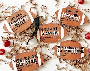 May include: Valentine's Day cards shaped like brown American footballs, with white stripes and messages. Phrases include "We are friendship goals," and "You're an all-star." Gold confetti and red sweets are scattered around the cards.