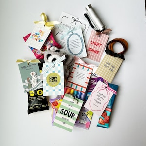 May include: A collection of gift items, including tags with inspirational quotes, a pen, hair ties, and small treats. The tags feature various designs and colors, tied with ribbons. The items are arranged on a white surface.