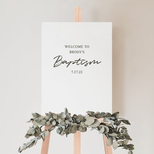 May include: White sign with black text on an easel. The sign reads "Welcome to Brody's Baptism 7.17.21".