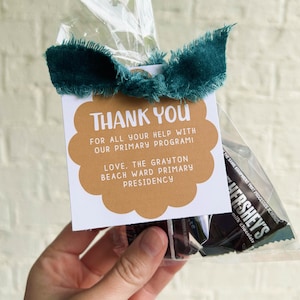 Pode incluir: Uma embalagem transparente de celofane amarrada com uma fita verde-azulado, contendo barras de chocolate ao leite Hershey's e uma etiqueta de presente. A etiqueta tem formato de vieira marrom claro com as palavras "THANK YOU" e uma mensagem.