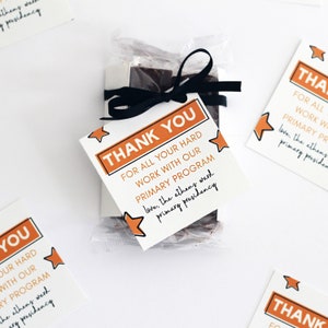 Könnte beinhalten: Eine kleine Geschenktüte mit einer Schokoladentafel darin. Die Tüte ist mit einem schwarzen Band gebunden und hat einen Dankesanhänger. Der Anhänger ist orange mit schwarzem Text und einem Sternendesign. Der Text auf dem Anhänger lautet "Vielen Dank für Ihre harte Arbeit mit unserem Primarprogramm. Liebe Grüße, die Primarpräsidentschaft des Athener Bezirks."