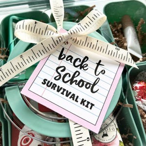 May include: A teal-colored container filled with various items, tied with a cream-colored ribbon printed with measurement markings. A tag with the words "back to School SURVIVAL KIT" is attached. The kit includes a lip balm and other small items.