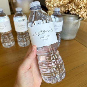 May include: A clear plastic water bottle with a white label featuring the black text "weston's BAPTISM 8.13.27" in a script and sans-serif font. The bottle has a ribbed design and a white cap, with additional identical bottles visible in the background.