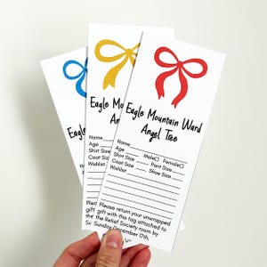 May include: Three white gift tags with colorful bow illustrations in blue, yellow, and red. Each tag has the text "Eagle Mountain Ward Angel Tree" and spaces for name, age, and wish list. The tags are held by a hand.