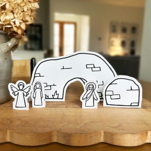 May include: A white nativity scene set with black outlines, including an angel, Mary, Joseph, and a stable. The figures are arranged on a wooden surface, with a blurred background featuring a vase of dried flowers.