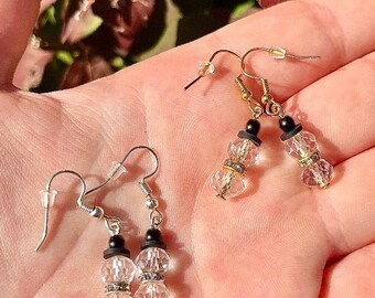 May include: Two pairs of snowman-shaped earrings. One pair has gold-colored hooks and accents, while the other has silver-colored hooks. Each snowman has a black top hat and clear, faceted crystal body with a decorative band.