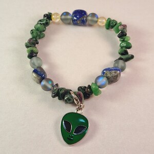 May include: A green gemstone bracelet with a silver charm of an alien face. The bracelet also features blue and gold beads.
