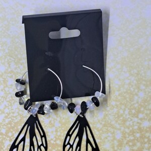 Könnte beinhalten: Ein Paar silberne Creolen mit schwarzen und weißen Edelstein-Akzenten und schwarzen Schmetterlingsanhängern, die unten hängen.