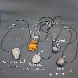 May include: Five necklaces with different gemstone pendants. The pendants are: Tiger's Eye, Merlinite, Tourmalinated Quartz, Rainbow Moonstone, and Star Ruby.