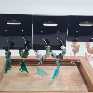 May include: Three pairs of hoop earrings with dinosaur charms. The earrings are made with various semi-precious stones, including green, blue, and pink.