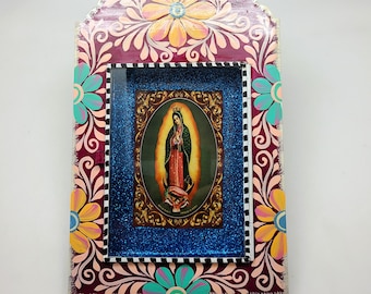 May include: A wooden frame with a colorful floral design surrounds a blue and silver glitter background with a picture of the Virgin Mary in a gold frame.