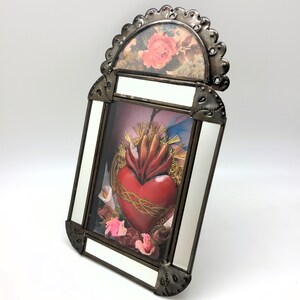 May include: A framed religious icon featuring a red heart with flames, a crown of thorns, and gold accents. The frame is metal with a floral print at the top and white borders. Pink and white flowers are at the base of the heart.