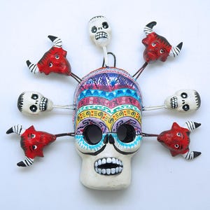 Op de afbeelding: Een kleurrijk Dia de los Muertos schedelmasker met zes kleinere schedels en duivelsfiguren eraan bevestigd. Het masker is geschilderd in felle kleuren, waaronder rood, geel, blauw en groen. De kleinere schedels zijn wit met zwarte ogen en monden.